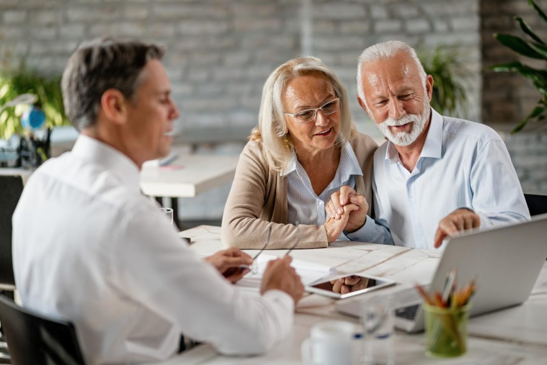 Happy Senior Couple Holding Hands Using Laptop While Having Meeting With Financial Advisor Office Senior Man Is Pointing Something Laptop E1686141134862